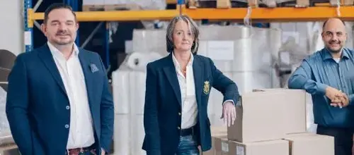 Das Team von Vogel Verpackungen steht im Lager zwischen Kartons auf Paletten
